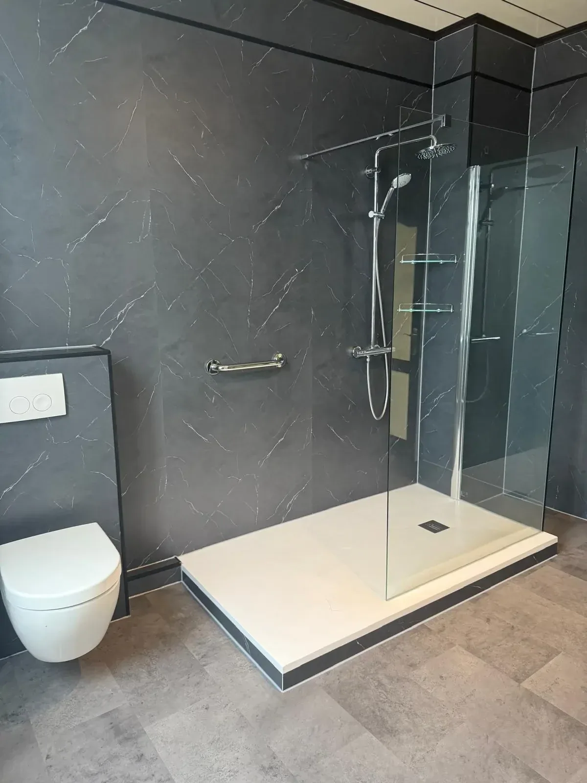 Badkamer met donkere marmerlook wandpanelen, inloopdouche met glazen wand, hangtoilet en steunbeugel