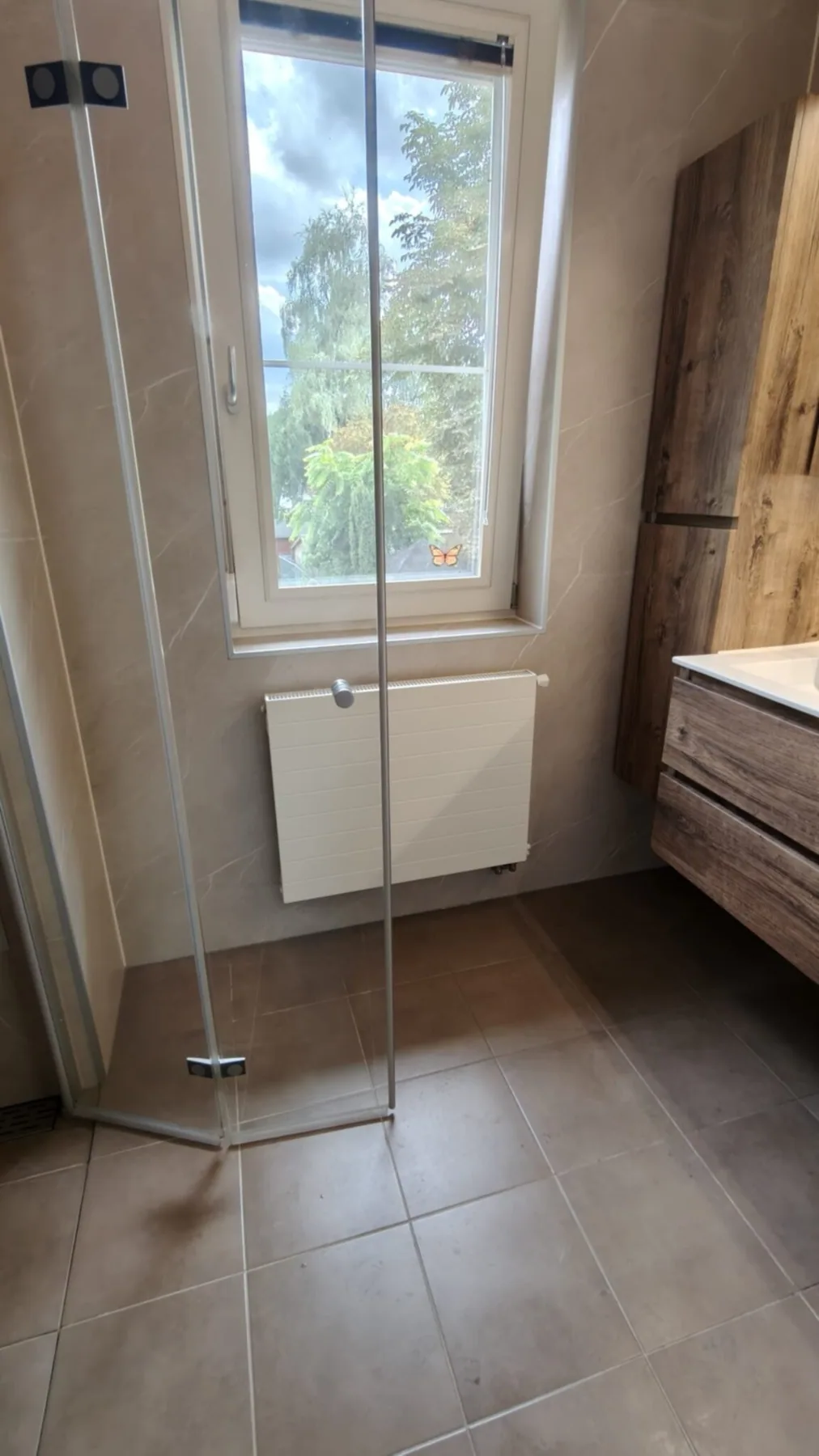 Glazen douchedeur voor raam met uitzicht op tuin, radiator en houtlook kast in lichte badkamer