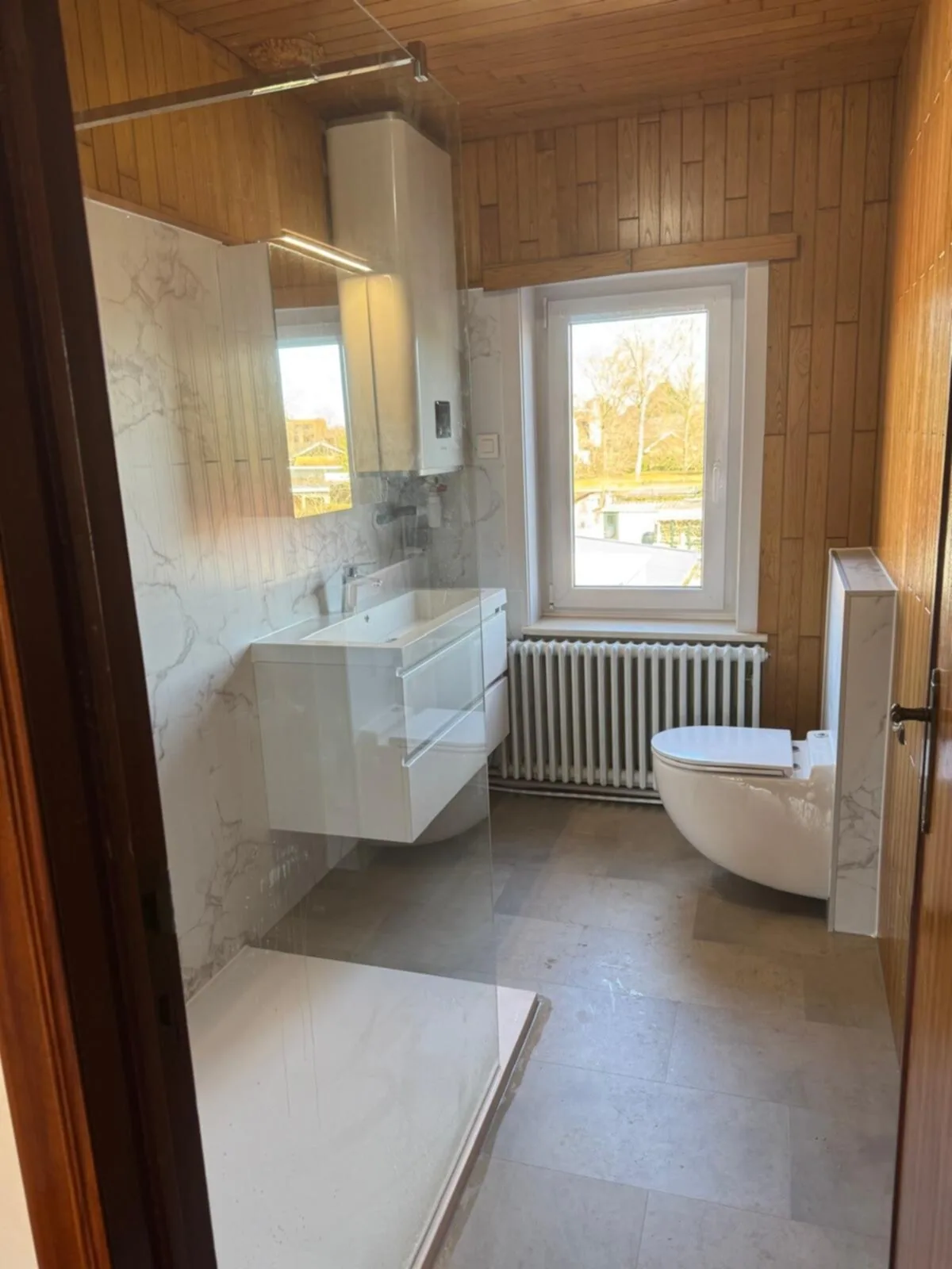 Badkamer met wit wastafelmeubel, hangtoilet, marmerlook wandpanelen en glazen douchewand onder houten plafond