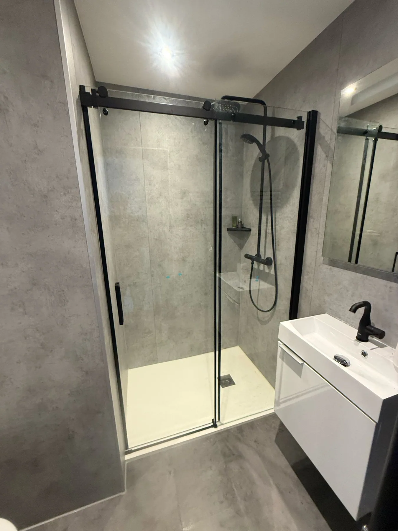 Douche met zwart omlijste glazen schuifdeur, betonlook wandpanelen, zwarte regendouche en wit wastafelmeubel
