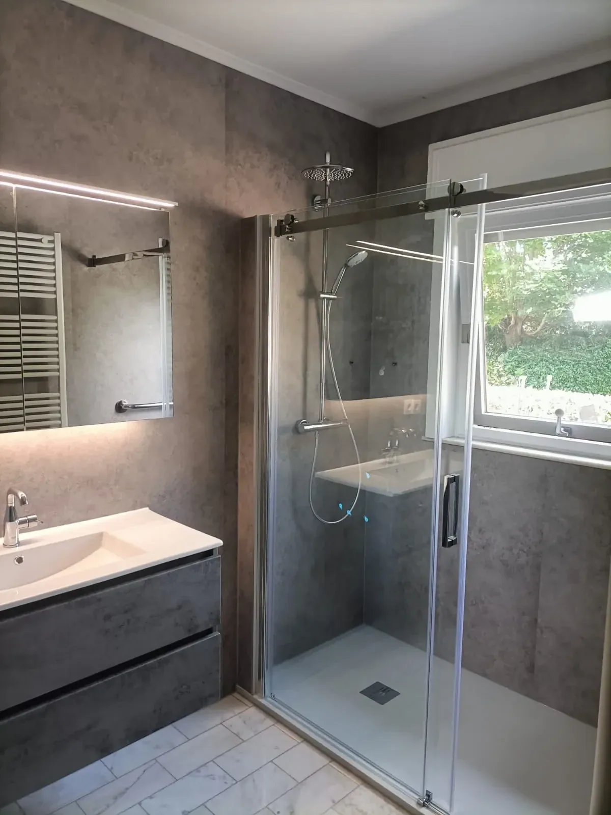 Badkamer met grijze steenlook panelen, douche met glazen schuifdeur, wastafelmeubel en verlichte spiegel