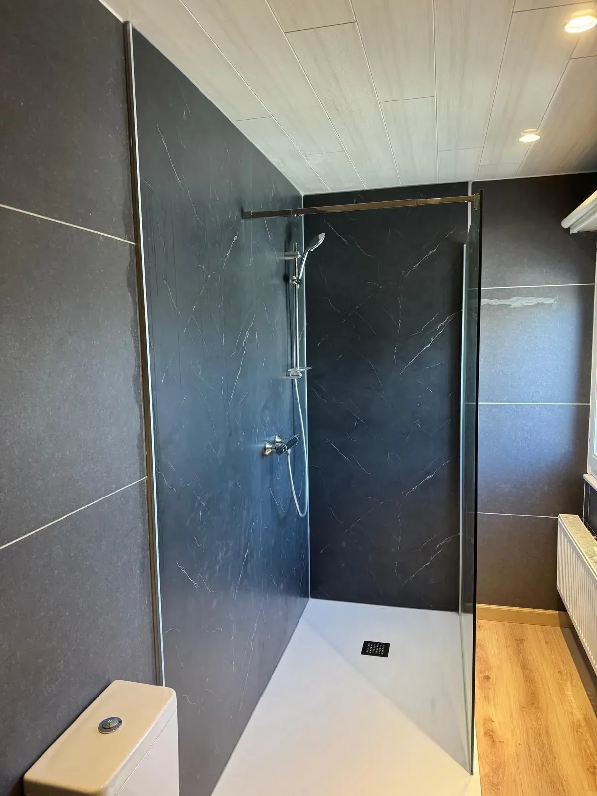 Inloopdouche met donkere marmerlook wandpanelen, glazen wand, houten vloer en staand toilet