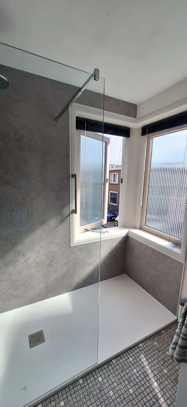 Ruime inloopdouche bij erkerraam met grijze betonlook wandpanelen, glazen wand en witte douchevloer