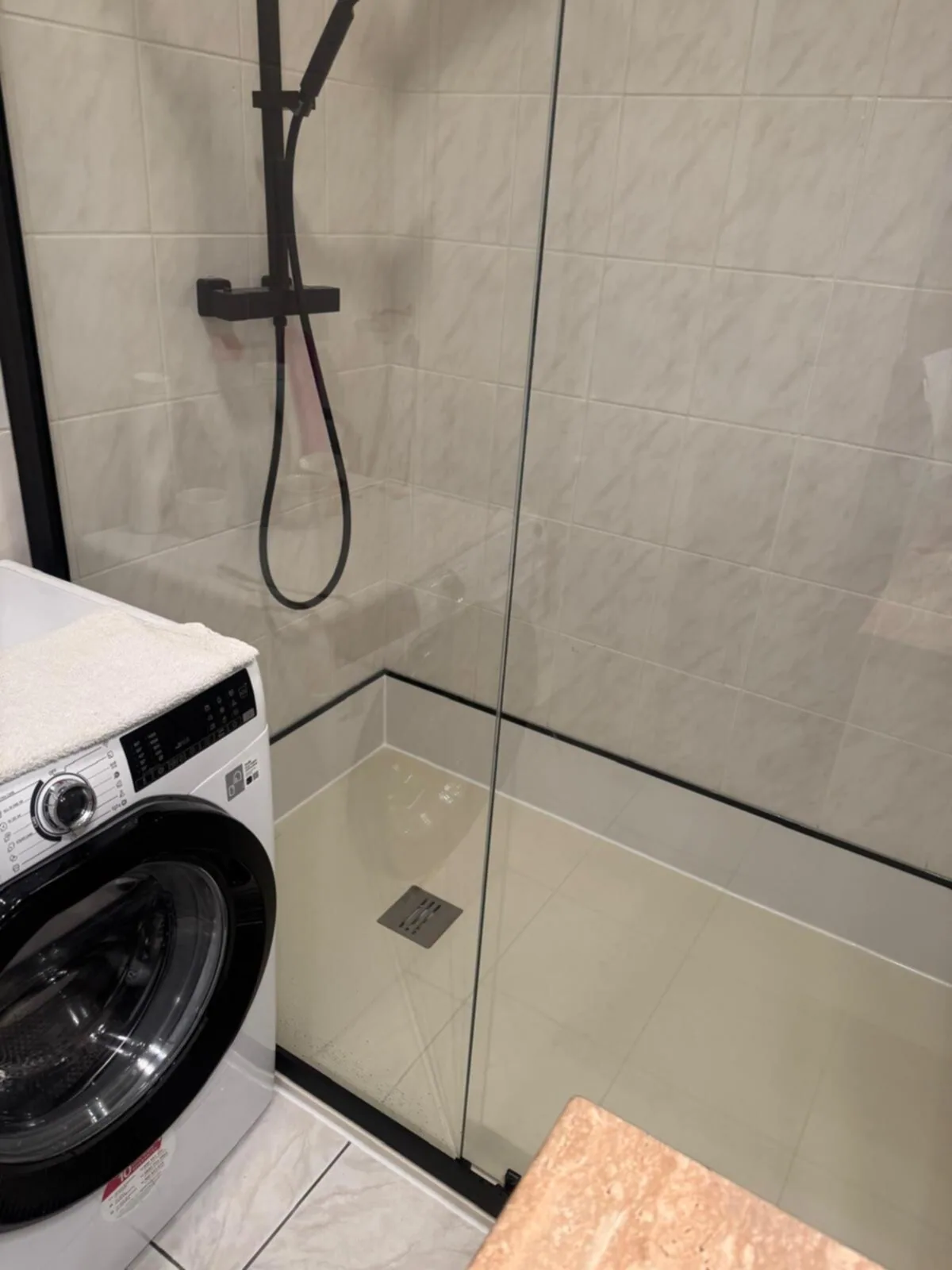 Inloopdouche met zwart omlijste glazen wand en zwarte thermostaatkraan naast wasmachine