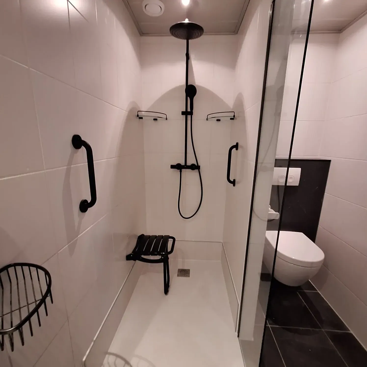 Witte inloopdouche met zwarte regendouche, zwarte steunbeugels, opklapbaar zitje en hangtoilet ernaast