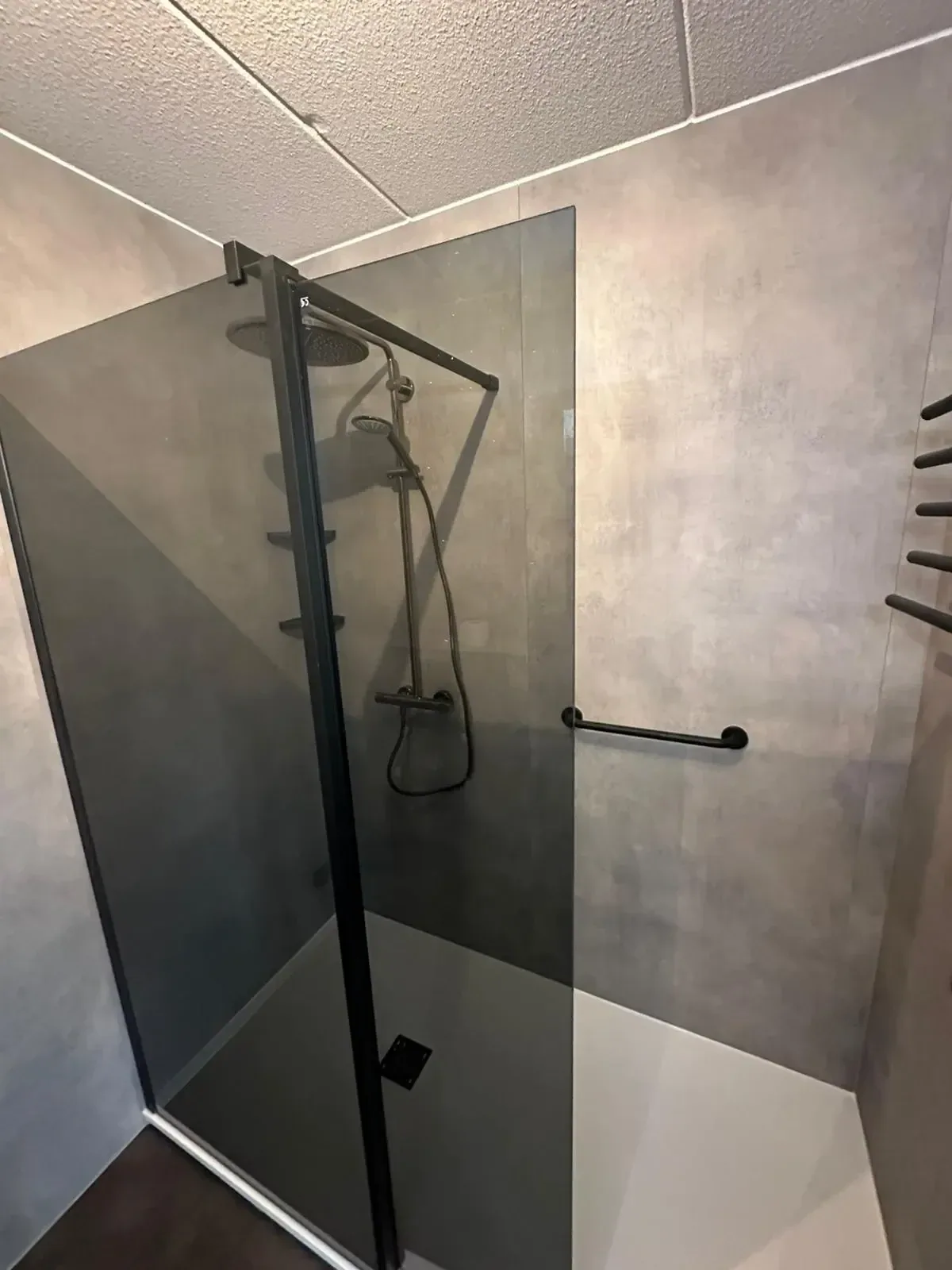 Rookglazen douchewand met zwart profiel, zwarte thermostaatkraan en steunbeugel tegen grijze wandpanelen