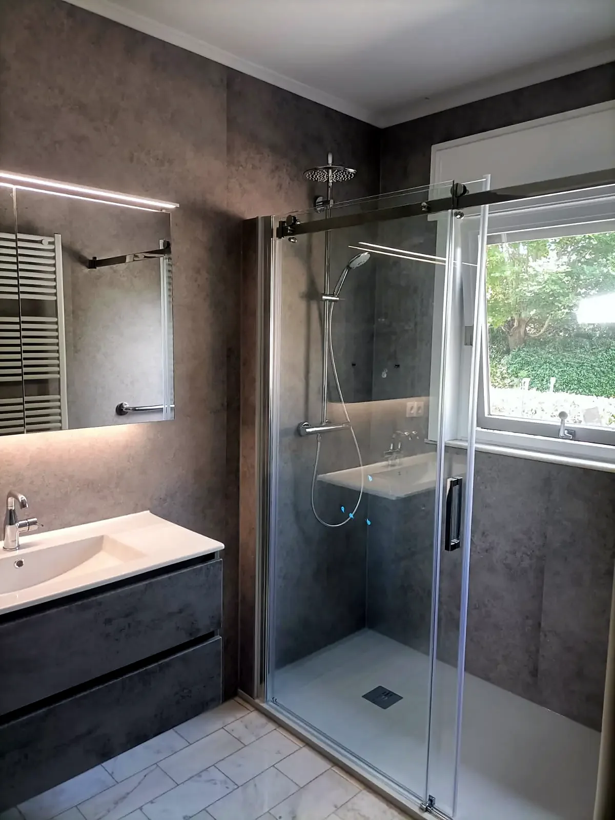 Douche met glazen schuifdeur naast een raam, grijs badmeubel met ledspiegel